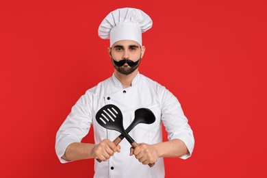 Photo of Professional chef with funny artificial moustache holding kitchen utensils on red background