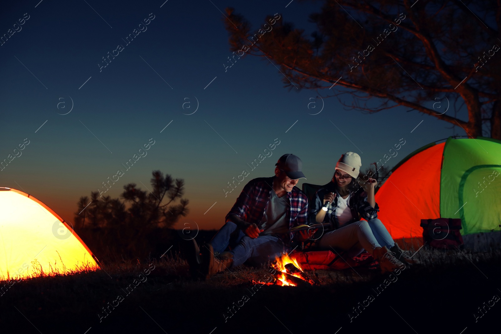 Photo of Couple with flashlight reading book near bonfire at night. Camping season