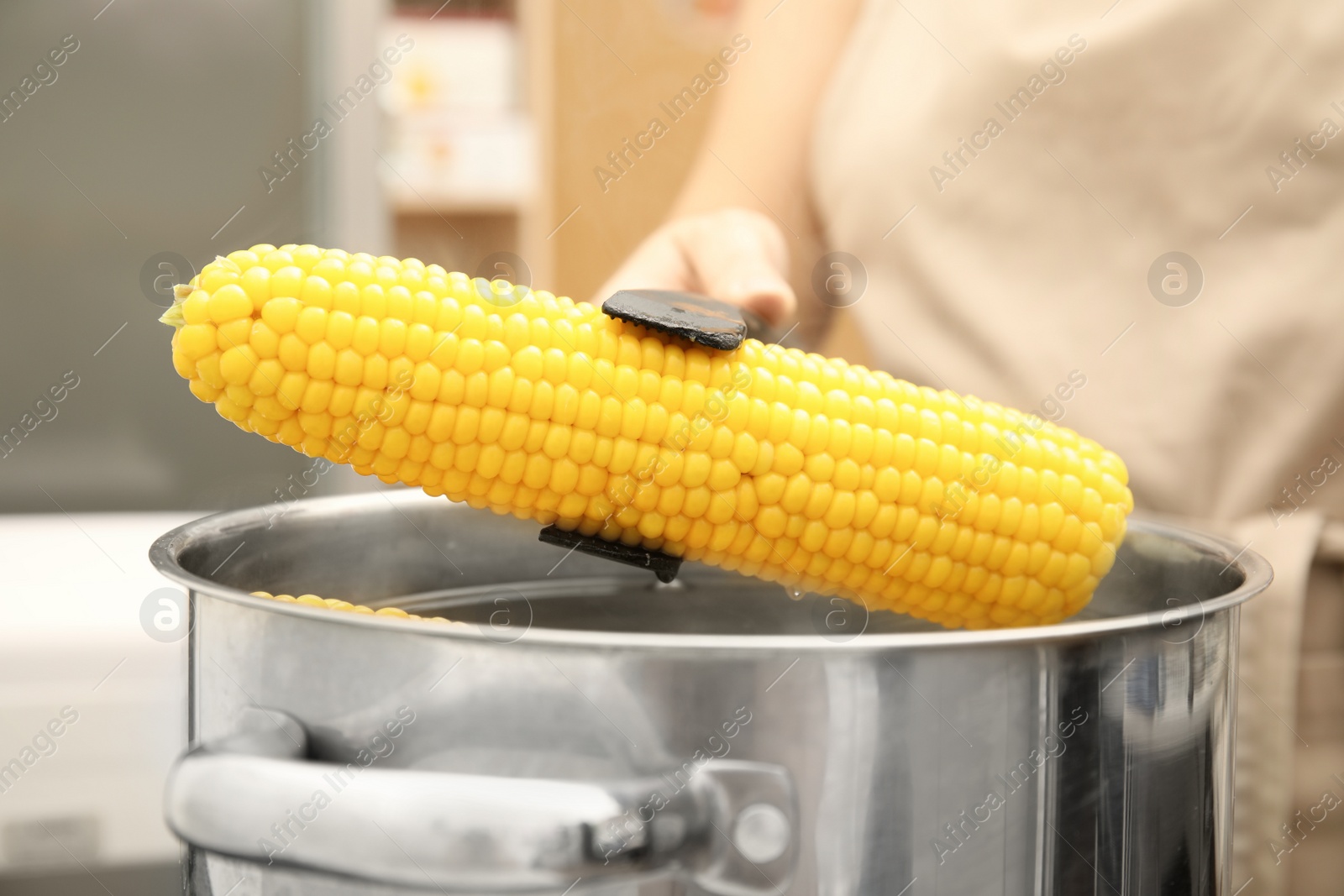 Photo of Woman taking boiled corn from pot with tongs in kitchen, closeup