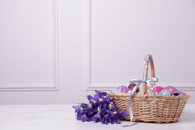Easter basket with painted eggs near bouquet of flowers on white marble table. Space for text