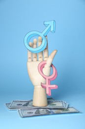 Gender pay gap. Wooden mannequin hand with symbols and dollar banknotes on light blue background