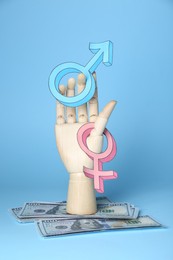 Photo of Gender pay gap. Wooden mannequin hand with symbols and dollar banknotes on light blue background