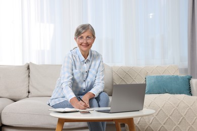 Photo of Beautiful senior woman writing something in notebook while working with laptop at home
