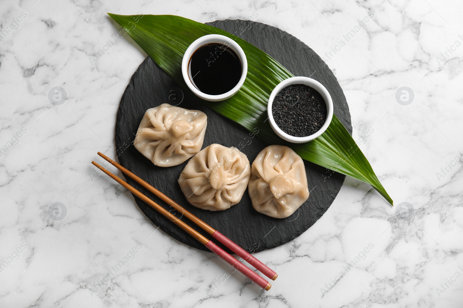 Photo of Slate plate with tasty baozi dumplings, chopsticks, sesame seeds and soy sauce on marble table, top view