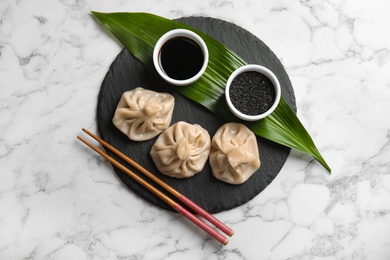 Photo of Slate plate with tasty baozi dumplings, chopsticks, sesame seeds and soy sauce on marble table, top view