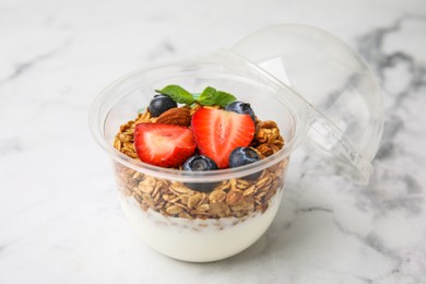 Photo of Tasty granola with berries and yogurt in plastic cup on white marble table, closeup