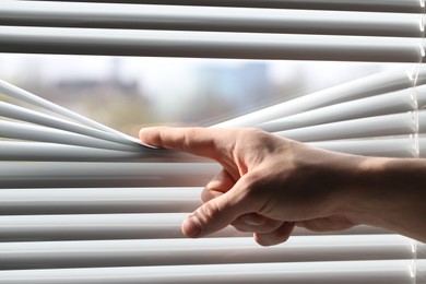 Man separating slats of white blinds indoors, closeup