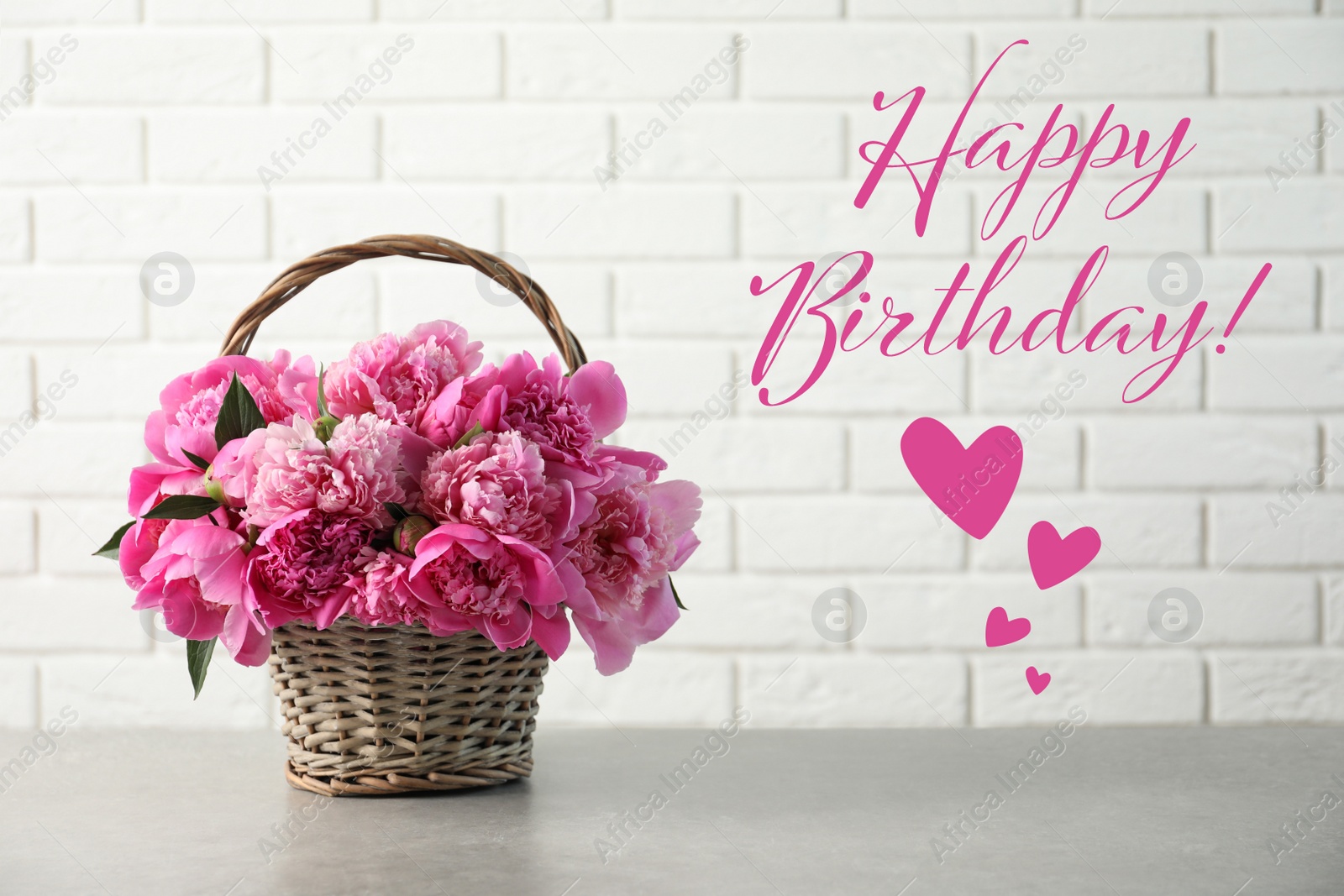 Image of Happy Birthday! Wicker basket with pink peonies on table near white brick wall
