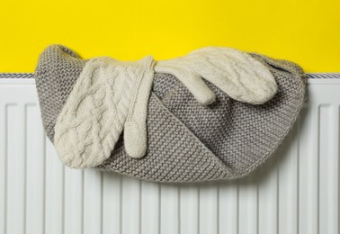 Photo of Knitted scarf and mittens on heating radiator near yellow wall