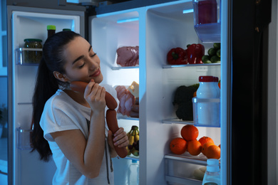 Photo of Young woman with sausages near open refrigerator at night