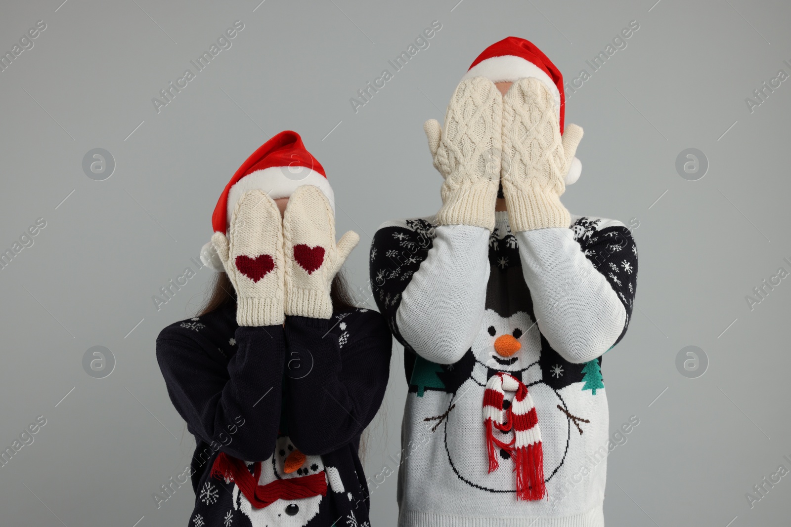 Photo of Young couple in Christmas sweaters and Santa hats covering faces with hands in knitted mittens on grey background