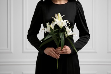 Woman with lilies near white wall, closeup. Funeral ceremony