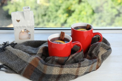 Photo of Cups of hot winter drink with scarf on window sill indoors