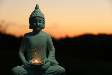 Decorative Buddha statue with burning candle outdoors at sunset. Space for text