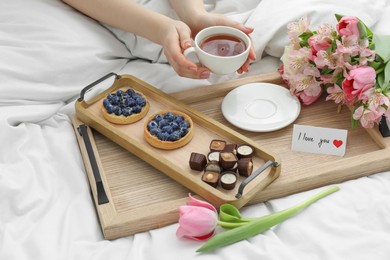 Photo of Tasty breakfast served in bed. Woman with tea, desserts, flowers and I Love You card at home, closeup