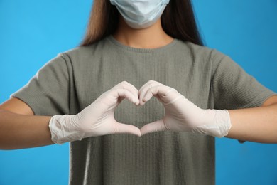 Photo of Woman in protective face mask and medical gloves making heart with hands on blue background, closeup