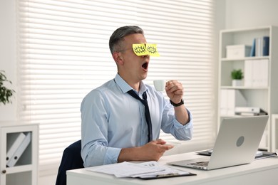 Man with fake eyes painted on sticky notes holding cup of drink and yawning at workplace in office
