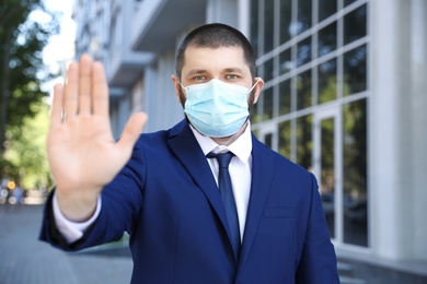 Photo of Man in protective face mask showing stop gesture outdoors. Prevent spreading of coronavirus