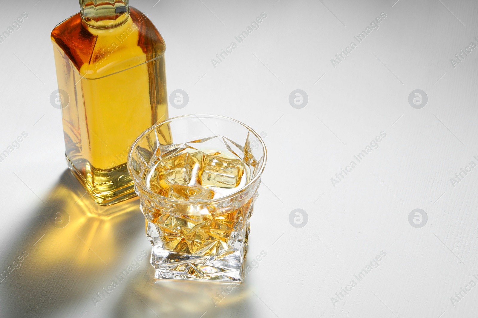 Photo of Whiskey with ice cubes in glass and bottle on white table, space for text