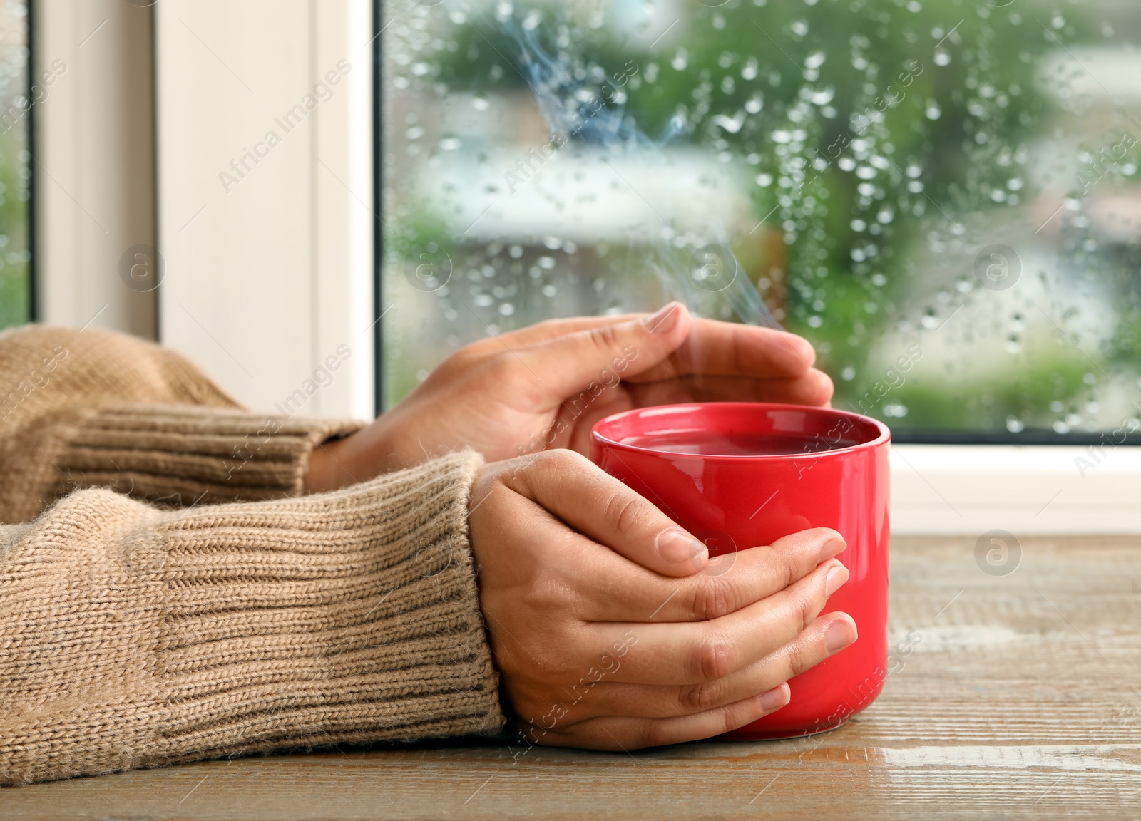 Photo of Woman with cup of hot drink at wooden table near window on rainy day, closeup