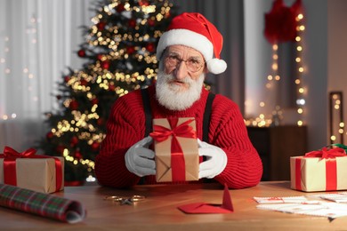 Photo of Santa Claus holding gift box at his workplace in room decorated for Christmas