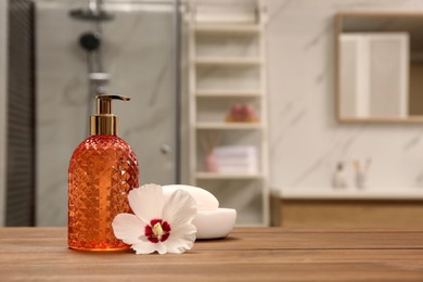 Photo of Glass dispenser with shower gel, soap bar and flower on wooden table in bathroom