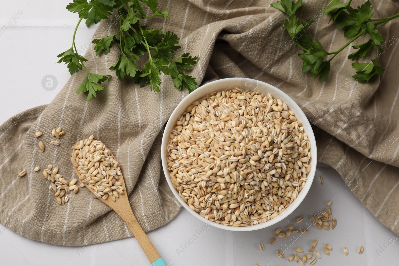 Photo of Dry pearl barley in bowl, wooden spoon and parsley on table, top view