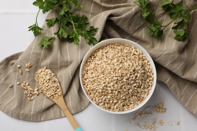 Photo of Dry pearl barley in bowl, wooden spoon and parsley on table, top view