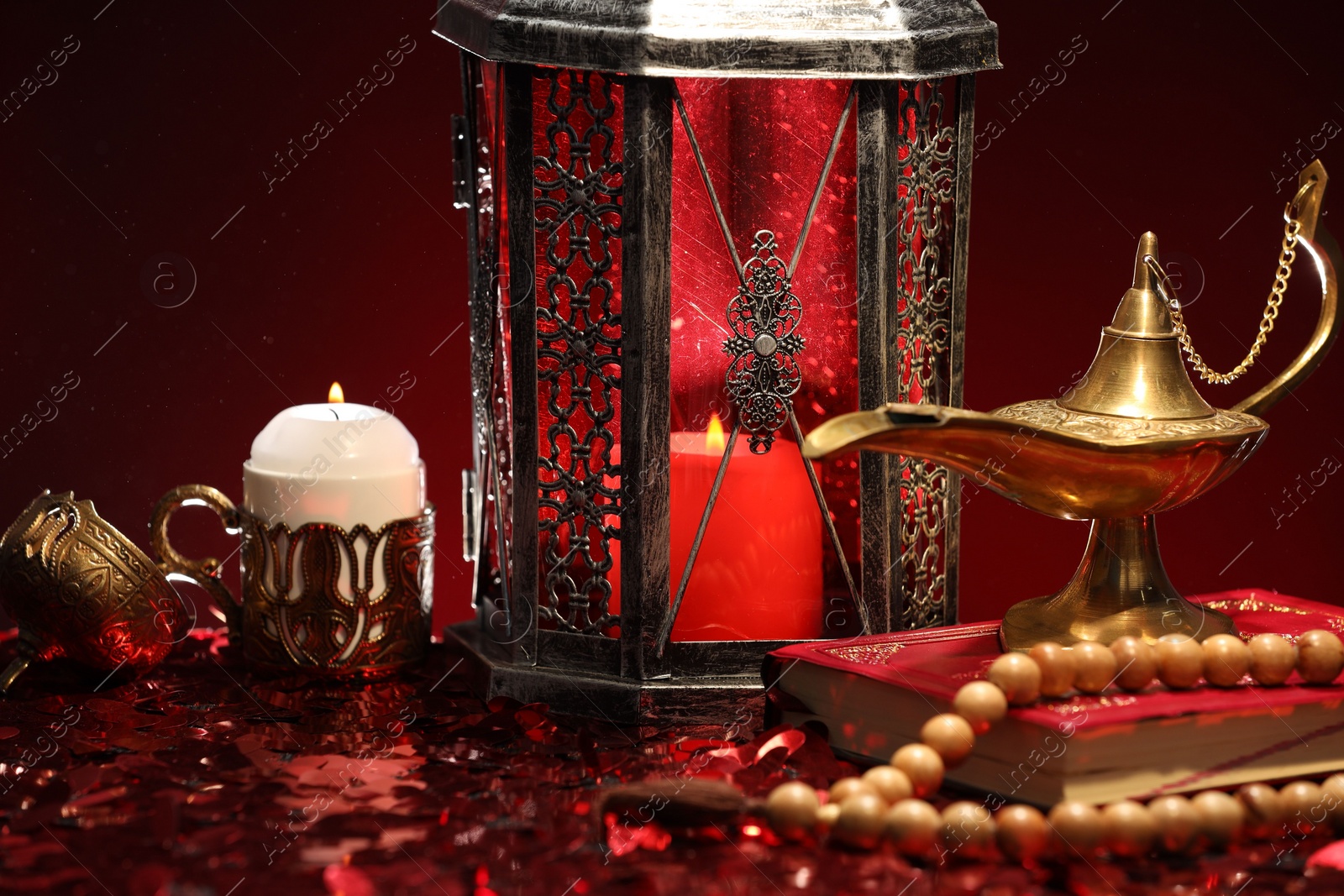 Photo of Arabic lantern, Quran, misbaha, burning candle and Aladdin magic lamp on shiny red table