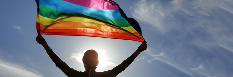 Image of Woman holding bright LGBT flag against blue sky on sunny day, back view. Banner design