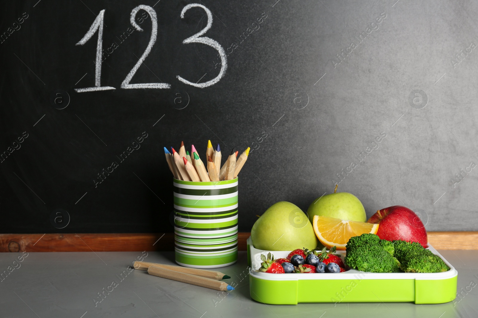 Photo of Healthy food for school child in lunch box and stationery on table near blackboard with chalk written numbers