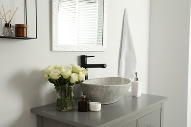 Vase with beautiful white roses and toiletries near sink in bathroom