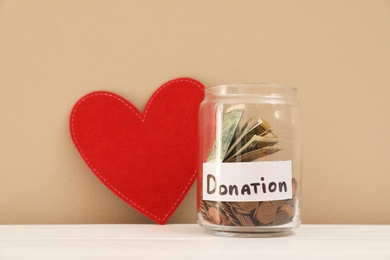 Photo of Red heart and donation jar with money on table against color background