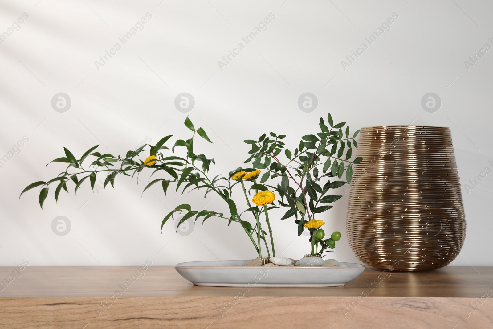 Photo of Stylish ikebana with beautiful yellow flowers, green branches and golden vase carrying cozy atmosphere at home