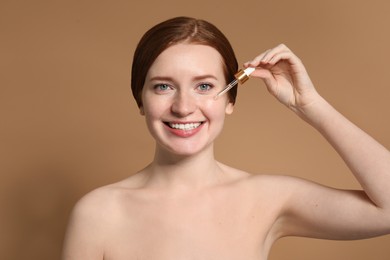 Smiling woman with freckles applying cosmetic serum onto her face on beige background