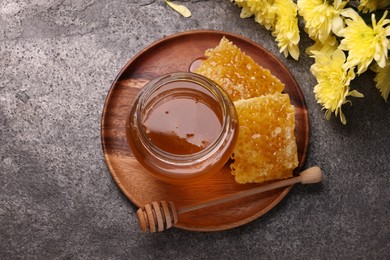 Photo of Sweet golden honey in jar, dipper, pieces of honeycomb and chrysanthemum flowers on grey textured table, flat lay