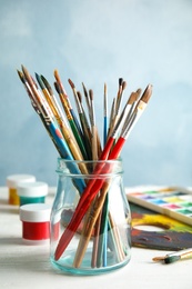 Glass jar with brushes and paints on table against color background