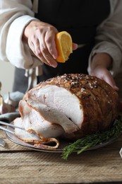 Photo of Woman squeezing juice from orange slice onto delicious baked ham at wooden table, closeup