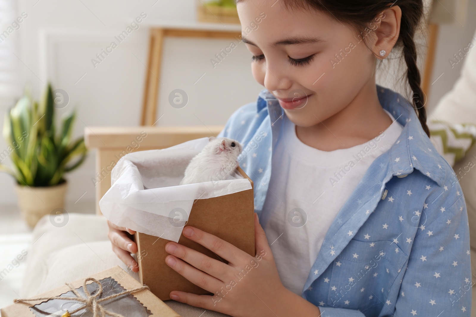 Photo of Happy little girl holding gift box with cute hamster at home