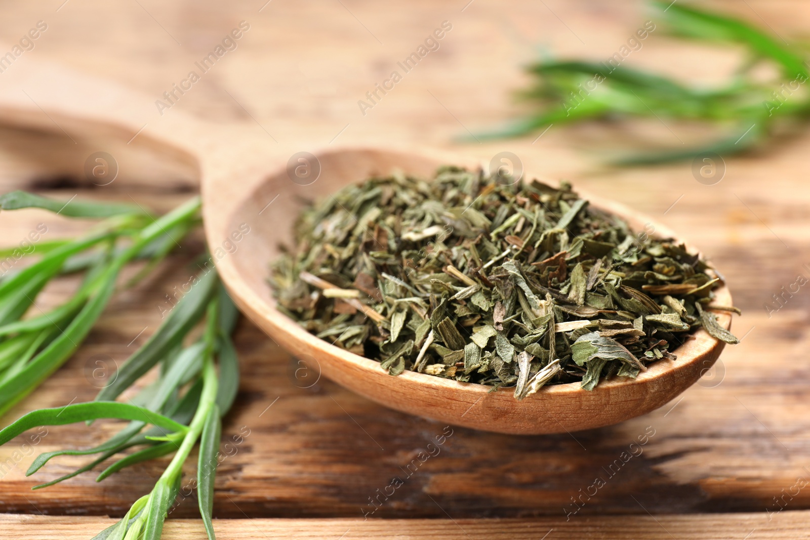 Photo of Spoon of dry tarragon and green leaves on wooden table, closeup