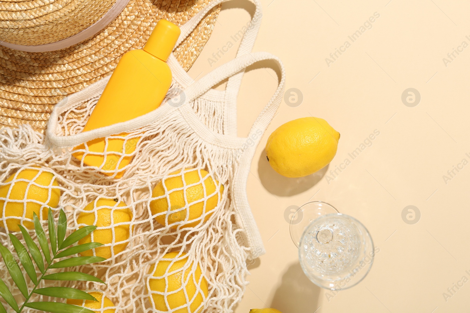 Photo of Fresh lemons, beach accessories in string bag and glass of drink on beige background, flat lay. Space for text