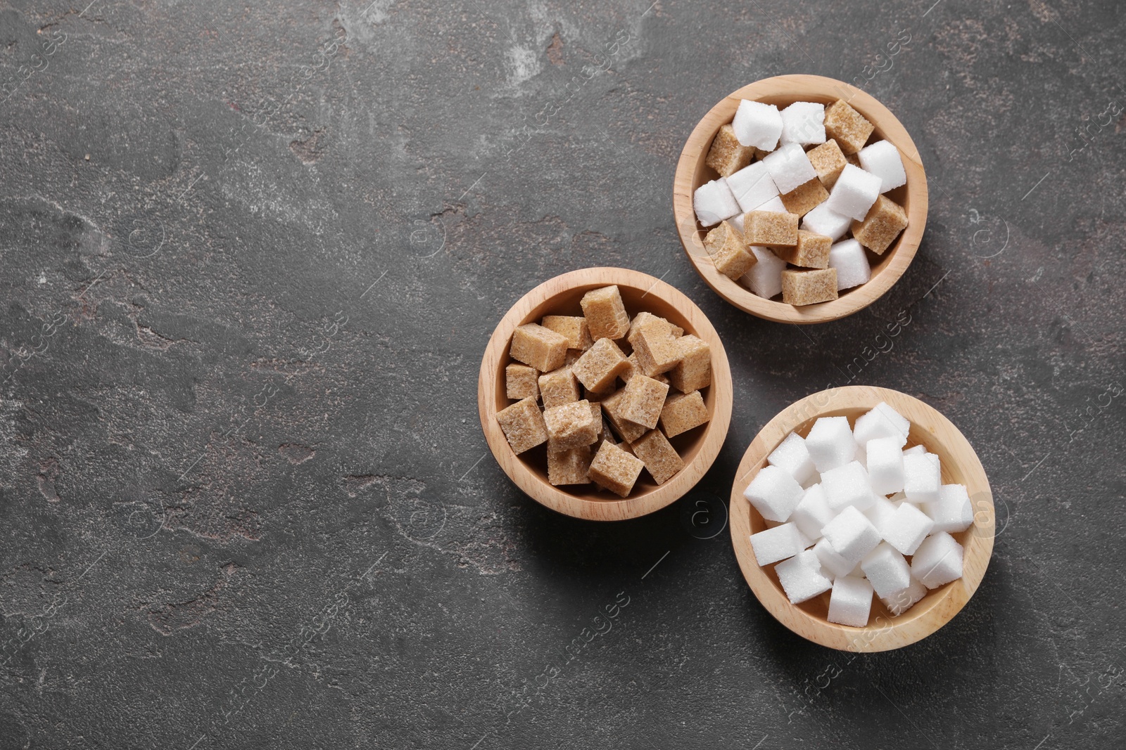 Photo of Different sugar cubes in bowls on gray textured table, flat lay. Space for text