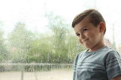 Cute little boy near window indoors, space for text. Rainy day