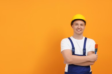 Happy worker holding paint brush near orange wall. Space for text