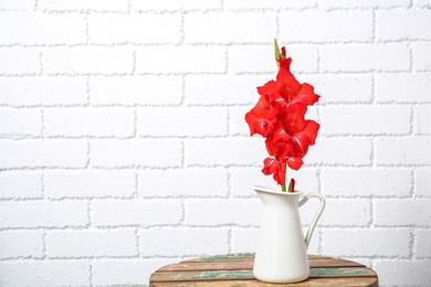 Photo of Jug with beautiful gladiolus flowers on table against brick wall