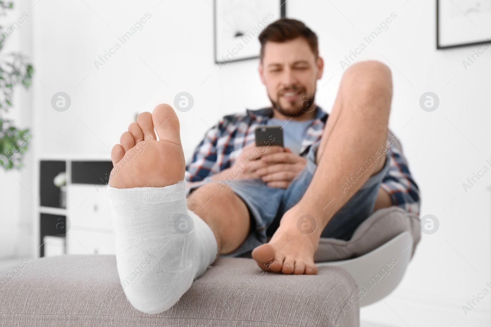 Photo of Man with broken leg in cast using mobile phone while sitting in armchair at home