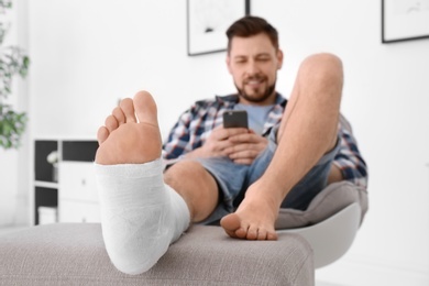 Man with broken leg in cast using mobile phone while sitting in armchair at home