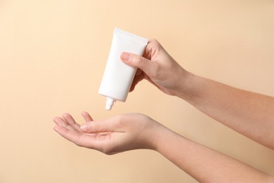 Woman applying cosmetic cream from tube onto her hand on beige background, closeup