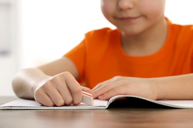 Photo of Little boy erasing mistake in his notebook at wooden desk, closeup