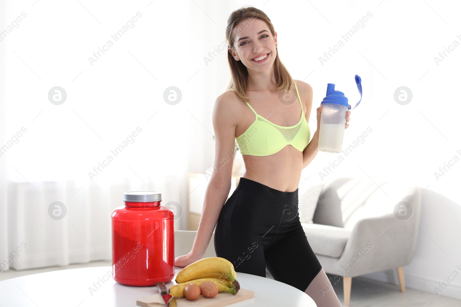 Photo of Young woman holding bottle of protein shake near table with ingredients in room. Space for text